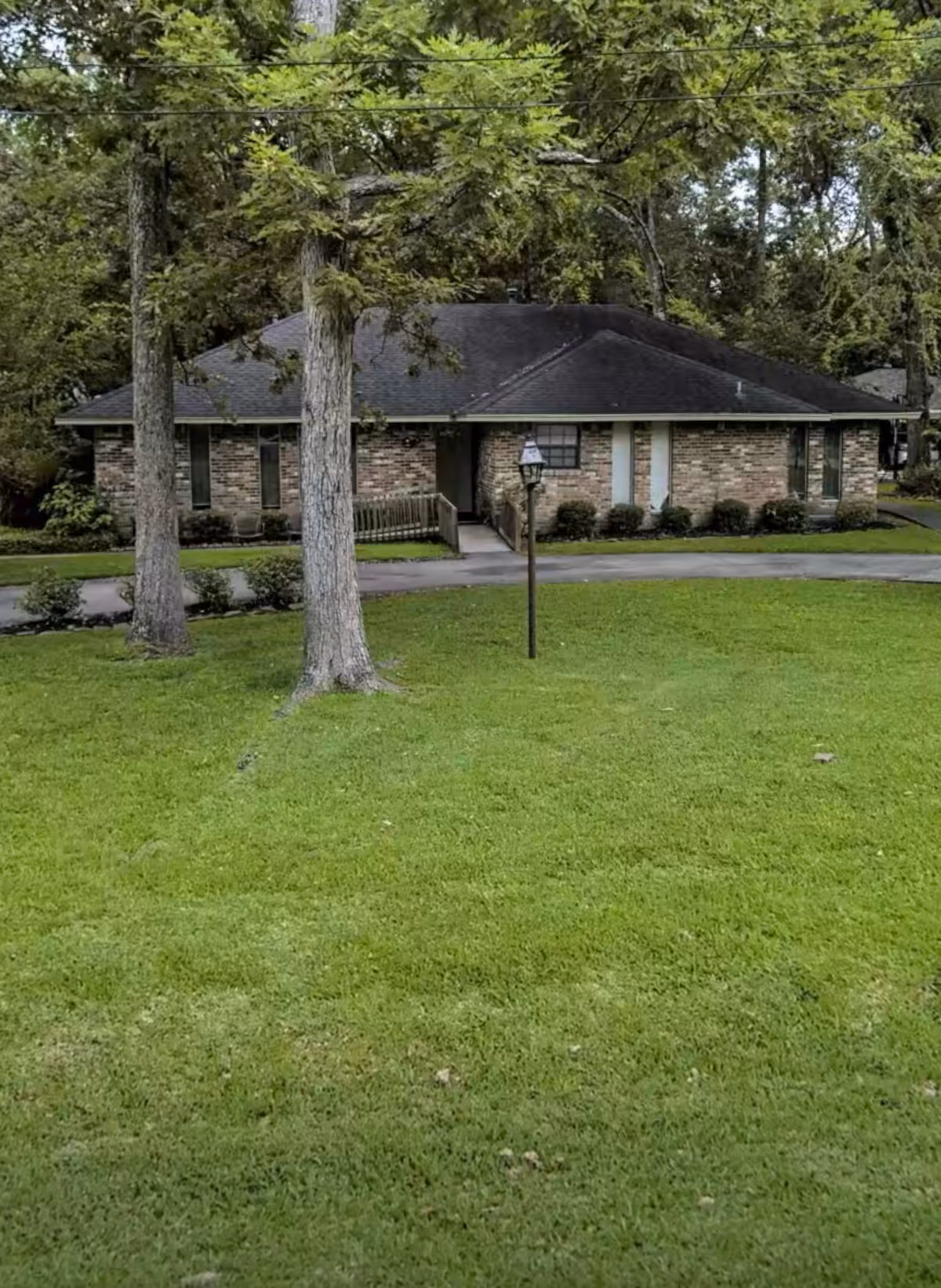 A single-story brick building with a dark shingled roof surrounded by trees and a well-maintained green lawn. A paved walkway leads to the entrance, and a lamp post stands near the path in front of the building.