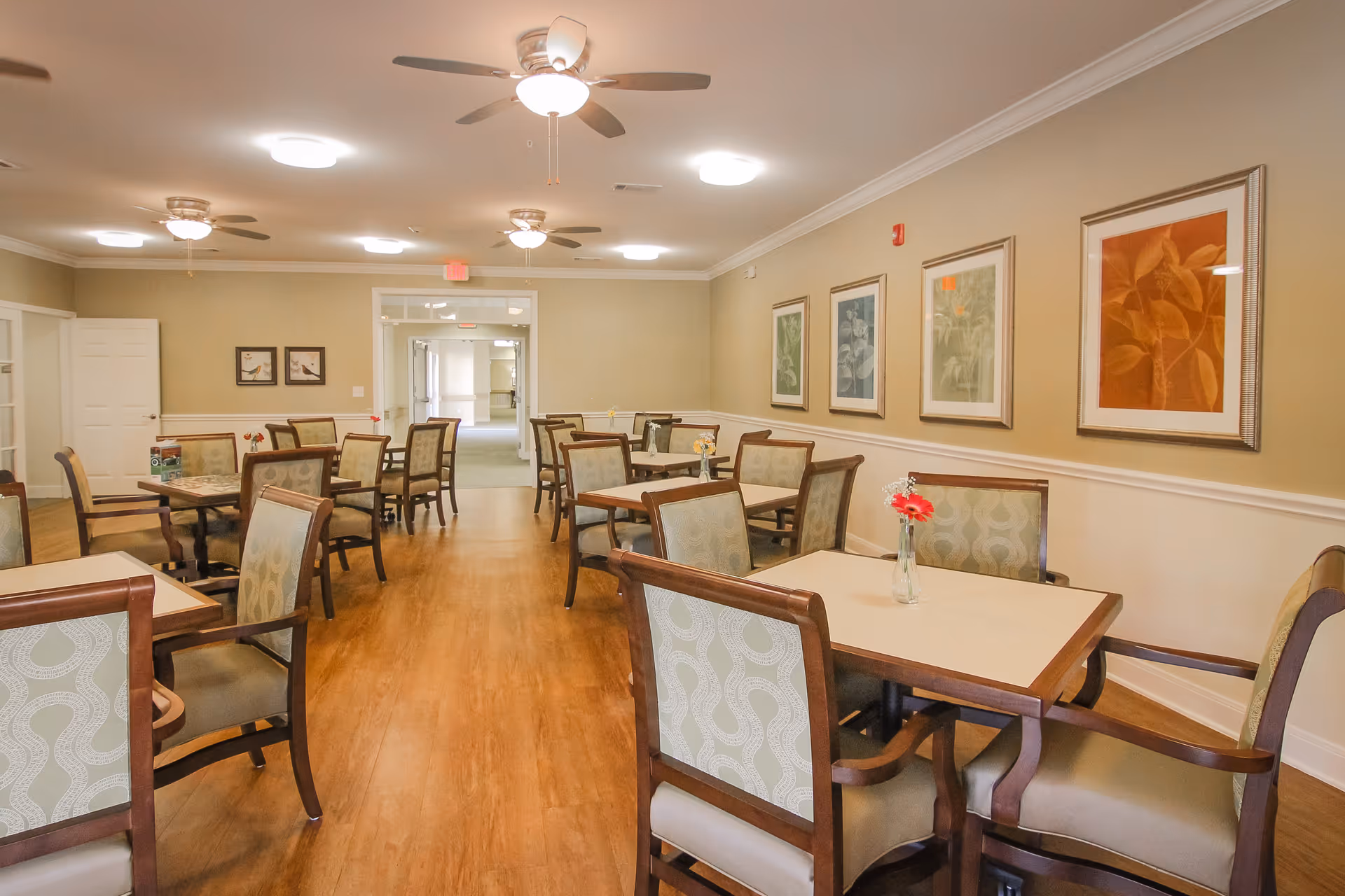 A dining room with multiple square tables and cushioned chairs arranged neatly on a wooden floor. The walls are beige with white trim and decorated with framed artwork. Ceiling fans and lights are mounted on the ceiling, and a hallway is visible in the background.