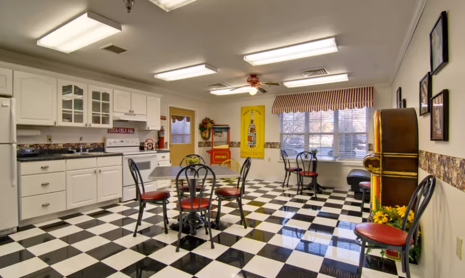 A retro-style dining area with a black and white checkered floor, white kitchen cabinets, a white refrigerator, stove, and sink. There are several black metal chairs with red cushions around small tables. A popcorn machine and a vintage Coca-Cola poster are visible near a window with striped valance curtains. The room is brightly lit with ceiling lights and a ceiling fan.