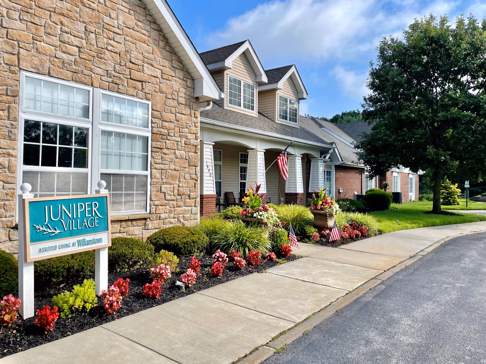 Exterior view of Juniper Village at Williamstown assisted living facility showing a stone and brick building with a porch, American flags, landscaped flower beds, and a sidewalk under a partly cloudy sky.