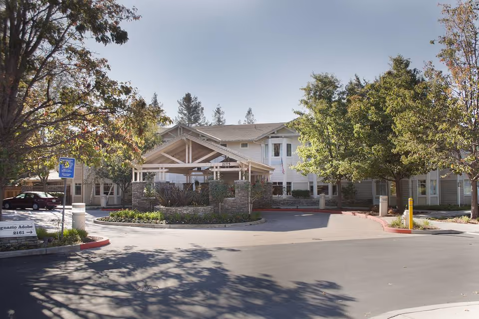 Street view of the front entrance of Ivy Park at Walnut Creek with a covered porte-cochere, landscaped roundabout and trees.