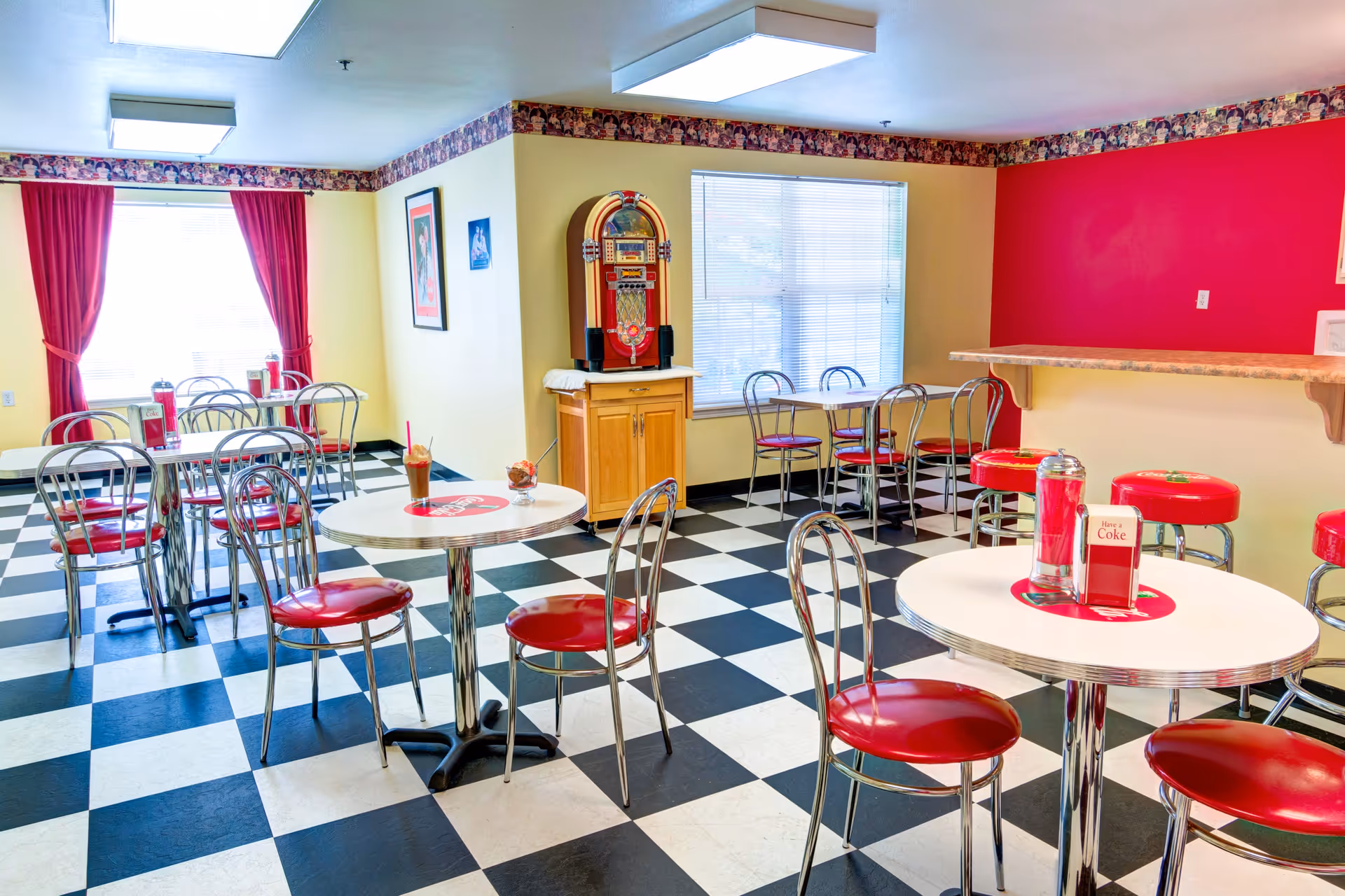 A retro-style dining area with black and white checkered floor tiles, red cushioned chairs, and round white tables. The room has yellow walls with a red accent wall and red curtains on the windows. There is a jukebox on a wooden cabinet and a counter with red bar stools.