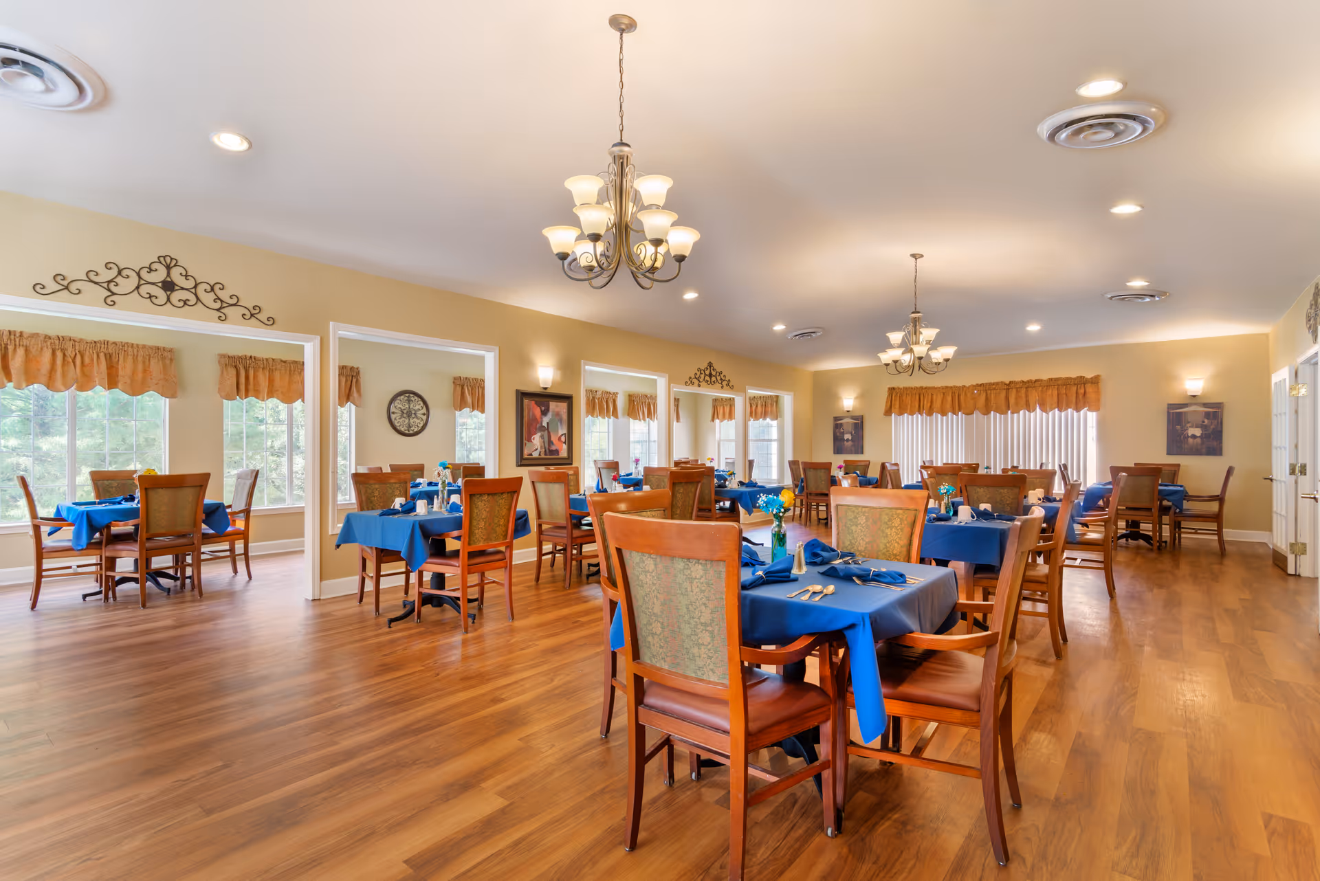 Spacious dining room with wooden floors, multiple tables set with blue tablecloths and chandeliers.