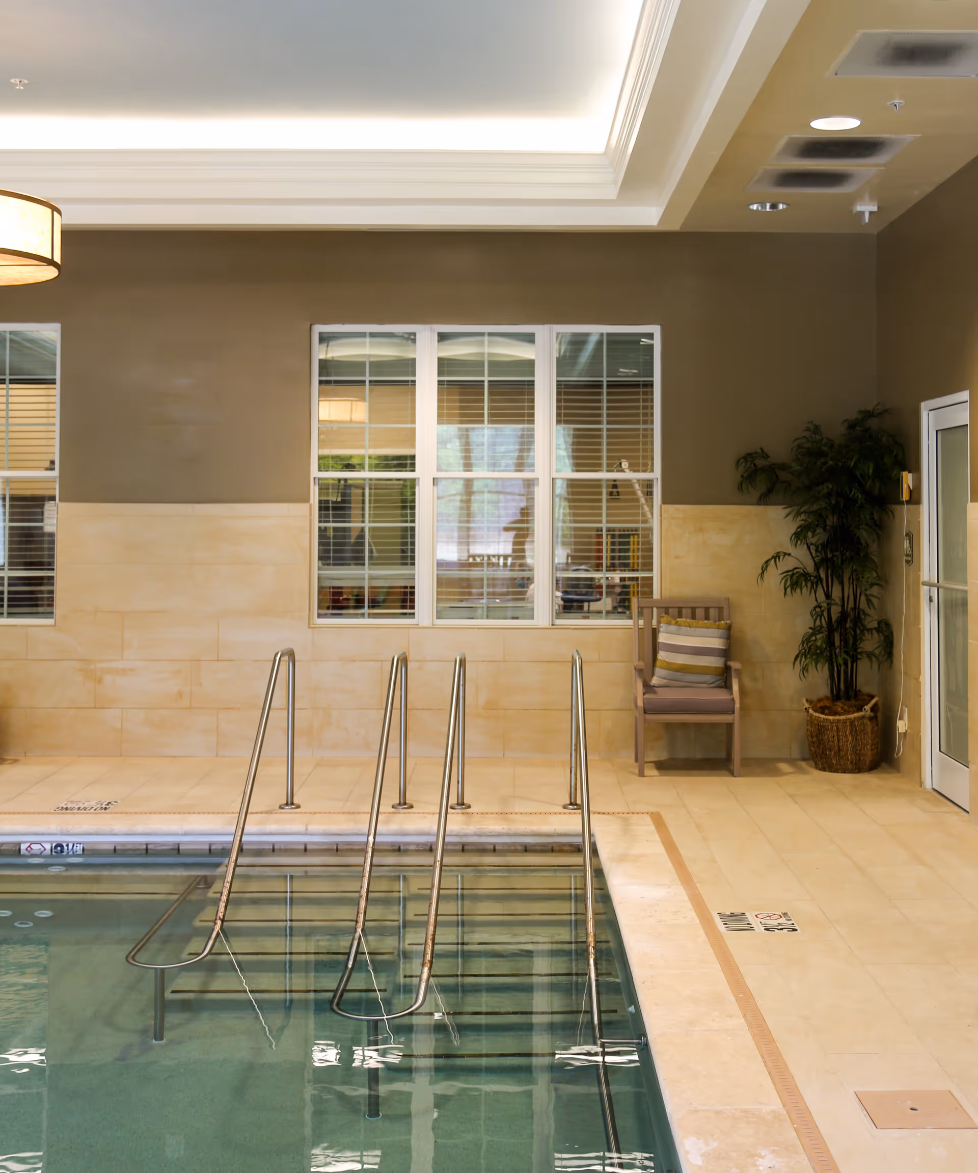 Indoor swimming pool with metal handrails leading into the water, beige tiled walls and floor, a wooden chair with a striped cushion, a potted plant, and windows with blinds on the wall behind the pool.