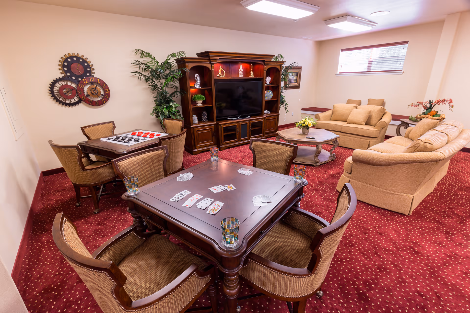 Carpeted common room with card tables and chairs, a TV cabinet, and sofas arranged for socializing.