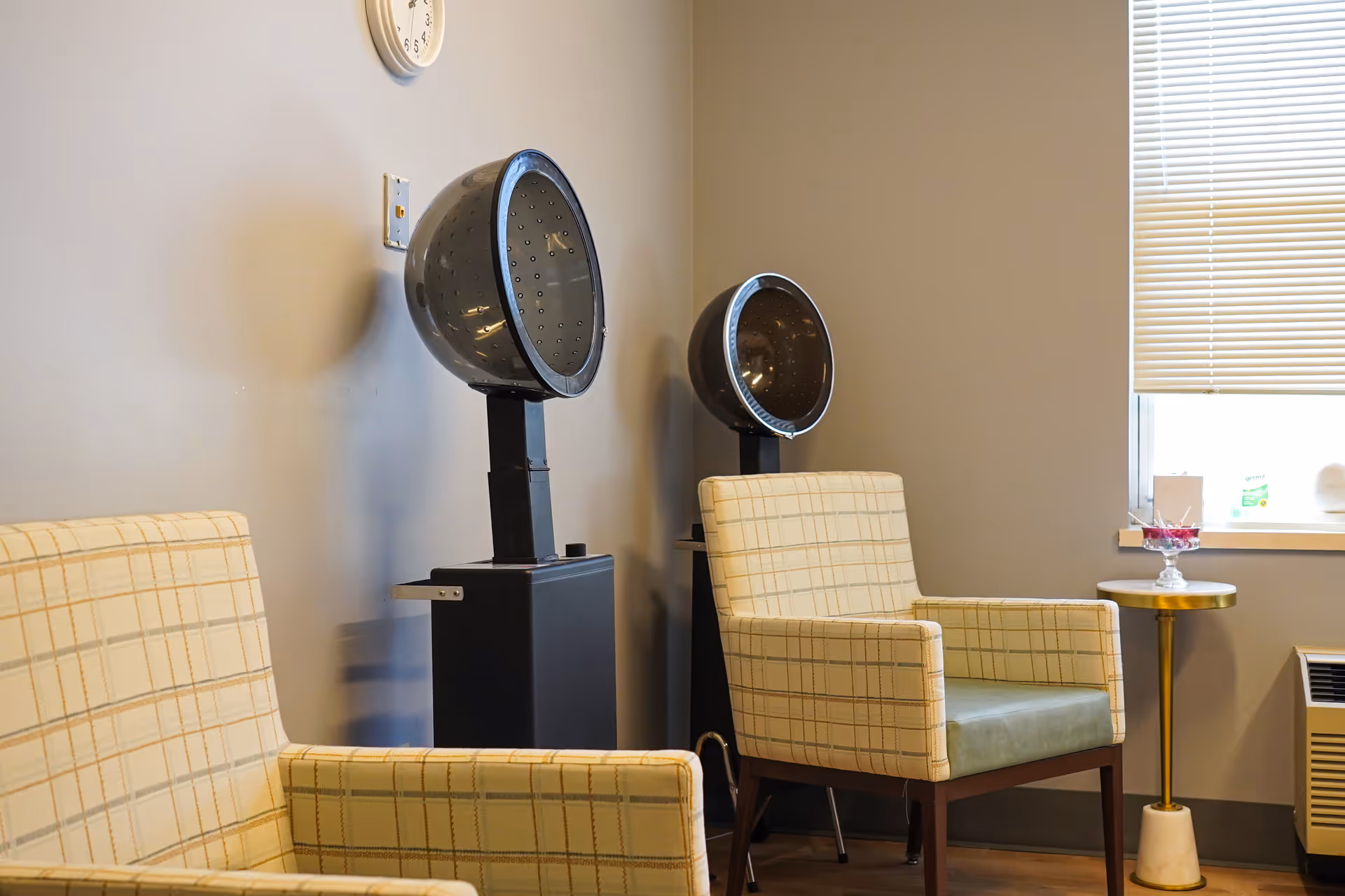 Interior seating area with two hooded hair dryers, two plaid armchairs, and a small side table beside a window.