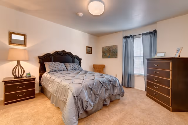 A bedroom with a large bed covered in a gray comforter, a wooden nightstand with a lamp and a small picture frame, a wooden dresser with framed photos on top, a window with blue curtains, a small orange chair in the corner, and beige carpeted floor.