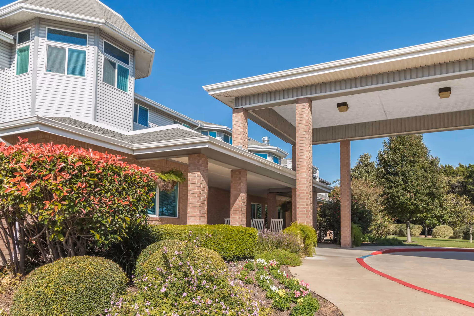 Exterior view of Asher Point Independent Living of Waco showing the building entrance with brick pillars, a covered driveway, well-maintained bushes, flowering plants, and trees under a clear blue sky.