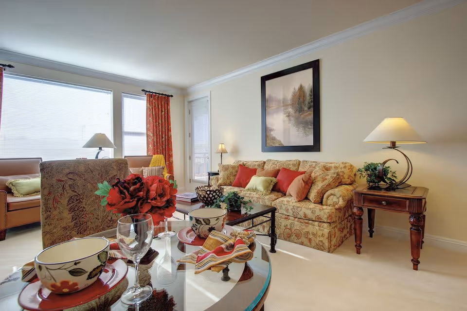 A cozy living room with a floral patterned sofa adorned with red and green cushions, a glass coffee table set with bowls, plates, and wine glasses, a side table with a lamp and plant, two armchairs near large windows with red curtains, and a framed landscape painting on the wall.