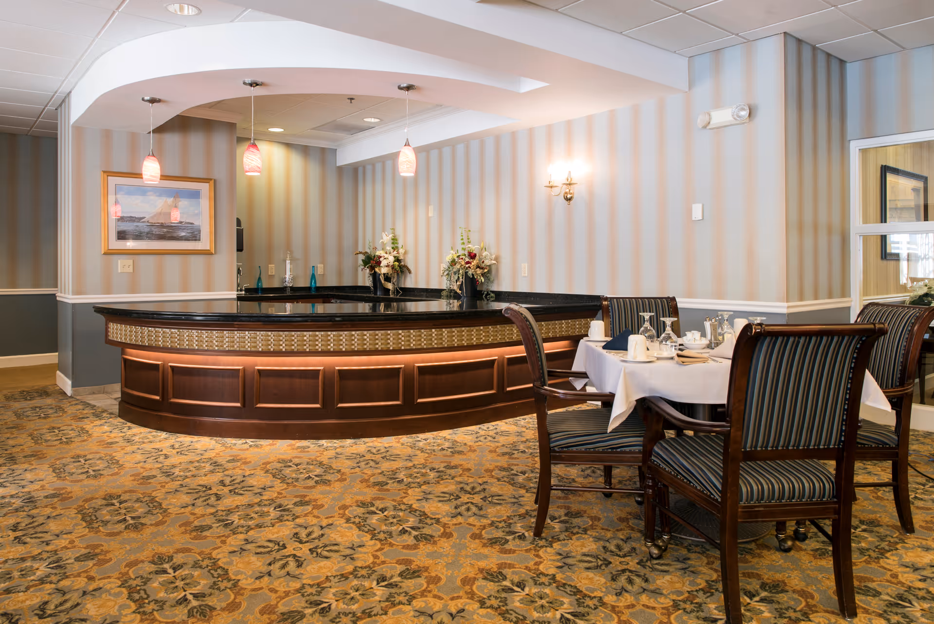 A dining area in a senior living facility featuring a wooden dining table set with white tablecloth, napkins, glassware, and silverware. The room has patterned carpet flooring, striped wallpaper, and a curved wooden counter with a black countertop and decorative lighting above. There are floral arrangements on the counter and a framed picture on the wall.