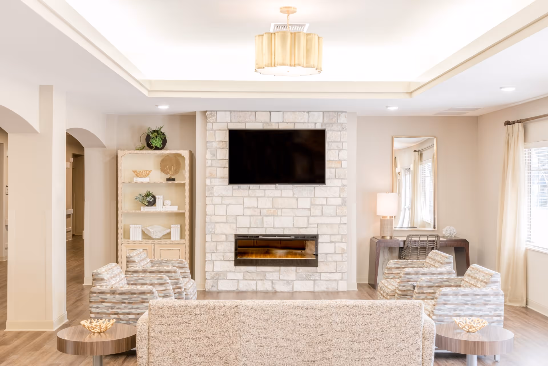 A bright and cozy living room with a stone fireplace and a mounted flat-screen TV above it. The room features a beige sofa facing the fireplace, four patterned armchairs arranged in pairs on either side, two round wooden side tables with decorative items, a tall bookshelf with plants and decor, a console table with a lamp and mirror, and large windows with light curtains allowing natural light to fill the space.