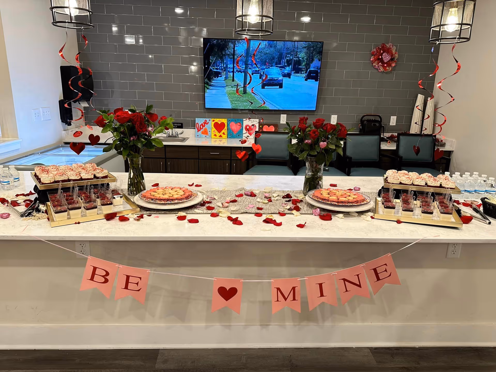 Valentine's-themed snack table in a senior living common area with roses, desserts, and a "Be Mine" banner.