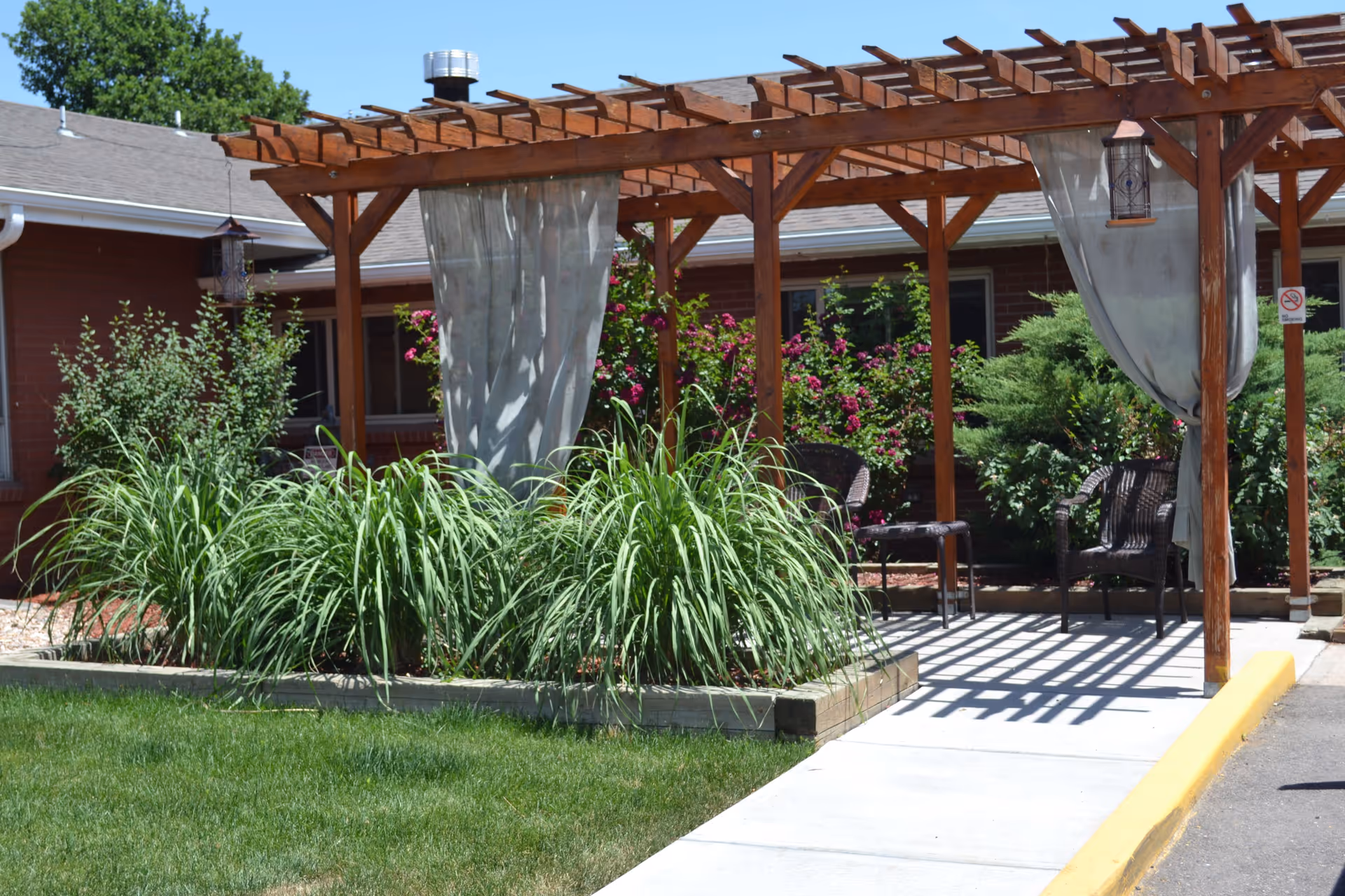 Outdoor seating area with a wooden pergola, light curtains hanging from the pergola, two wicker chairs, and lush green plants and bushes surrounding the space. The area is adjacent to a brick building with windows.