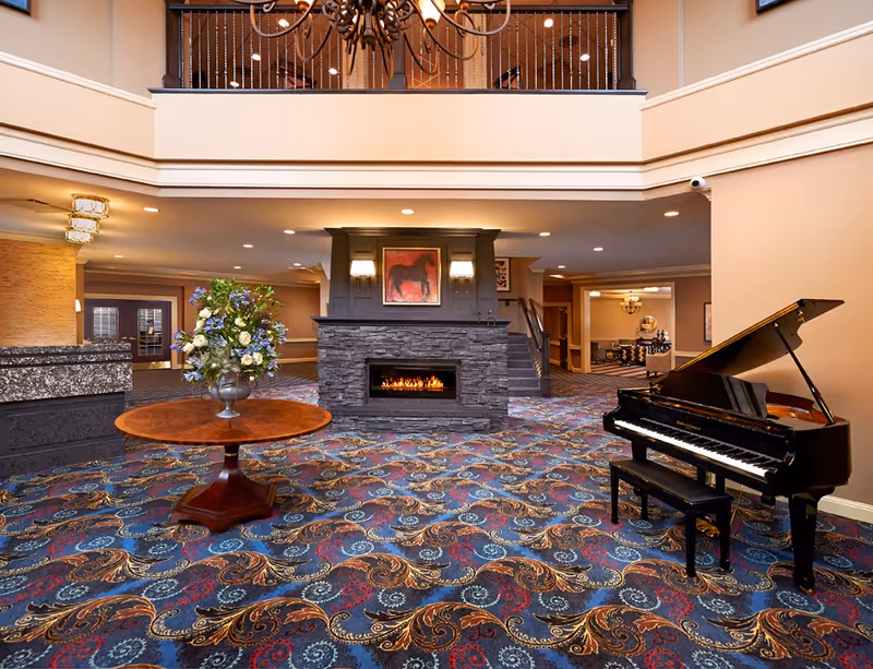 Spacious and elegant assisted living facility common area with a colorful patterned carpet, a round wooden table with a large floral arrangement, a black grand piano with a bench, a stone fireplace with a painting of a horse above it, and a balcony with a chandelier overhead.