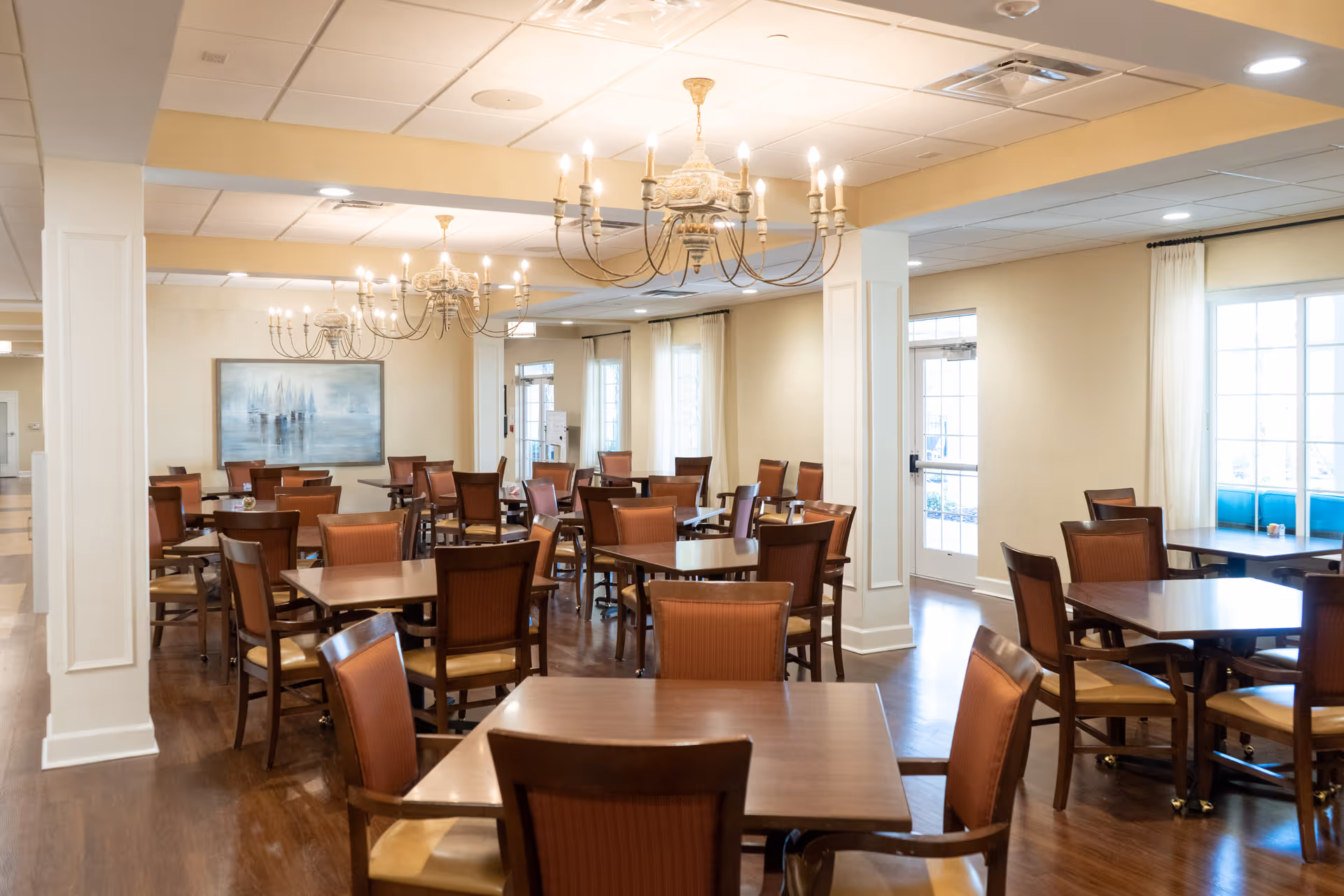 A spacious dining room with multiple wooden tables and chairs arranged neatly. The room features large windows with white curtains allowing natural light to fill the space. Elegant chandeliers hang from the ceiling, and a large painting is visible on one wall. The floor is wooden, and the overall ambiance is warm and inviting.