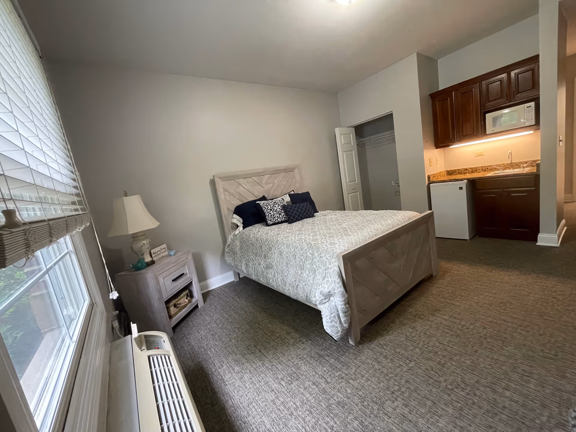 A cozy bedroom with a bed featuring a light wood headboard and footboard, decorated with patterned bedding and pillows. Next to the bed is a nightstand with a lamp and small decorative items. There is a window with blinds on the left side, and a small kitchenette with dark wood cabinets, a microwave, a mini fridge, and a sink on the right. An open closet space is visible near the kitchenette.
