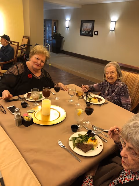 Three elderly women sitting around a dining table in a senior living facility, enjoying a meal together with plates of food and glasses of wine and water. The table is set with a candle centerpiece and condiments. The room has warm lighting and a framed picture on the wall.