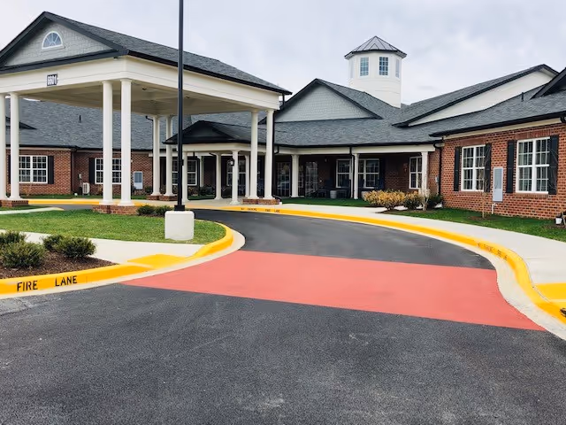 Exterior view of a senior living facility with a covered entrance driveway, red brick walls, multiple windows, and a cupola on the roof under a cloudy sky.