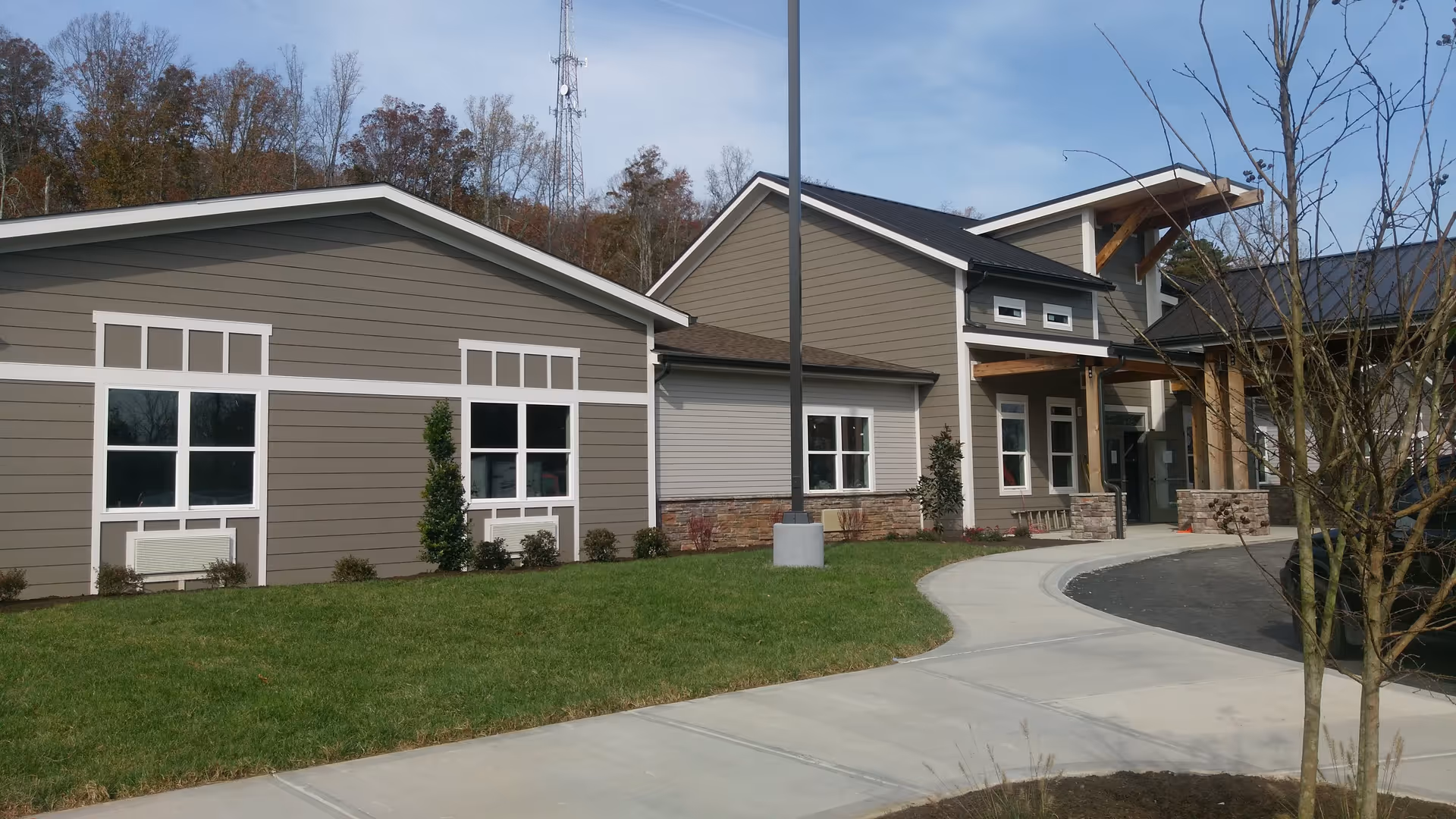 Exterior view of a modern assisted living facility building with beige and gray siding, white trim around windows, a covered entrance with wooden beams, a curved concrete walkway, green lawn, and some small trees and shrubs.
