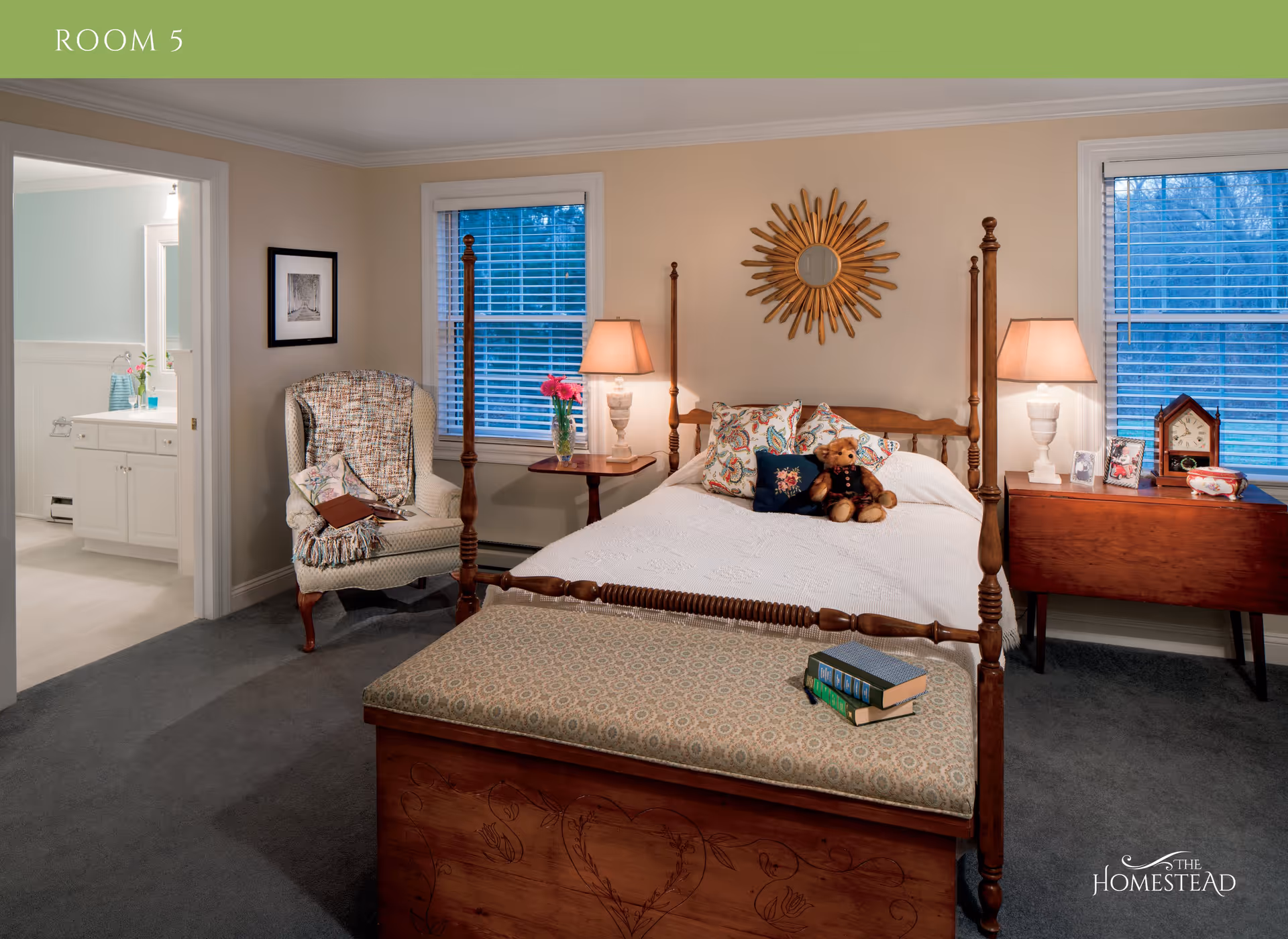 A cozy bedroom with a wooden four-poster bed adorned with patterned pillows and a teddy bear. At the foot of the bed is a cushioned bench with books on it. To the left, there is a comfortable armchair with a blanket and a book. Two windows with blinds flank the bed, and there are two bedside tables with lamps. A sunburst mirror hangs on the wall above the bed. An open door reveals a bathroom with a sink and mirror. The room has beige walls and gray carpet.