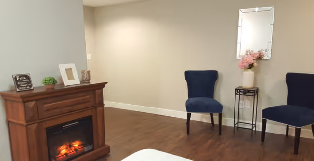 A cozy interior room with wooden flooring featuring a small electric fireplace cabinet on the left, decorated with a small plant, a framed quote, and a candle. Two navy blue upholstered chairs are placed against a beige wall, with a small black metal stand holding a vase of pink flowers and a rectangular mirror above it.