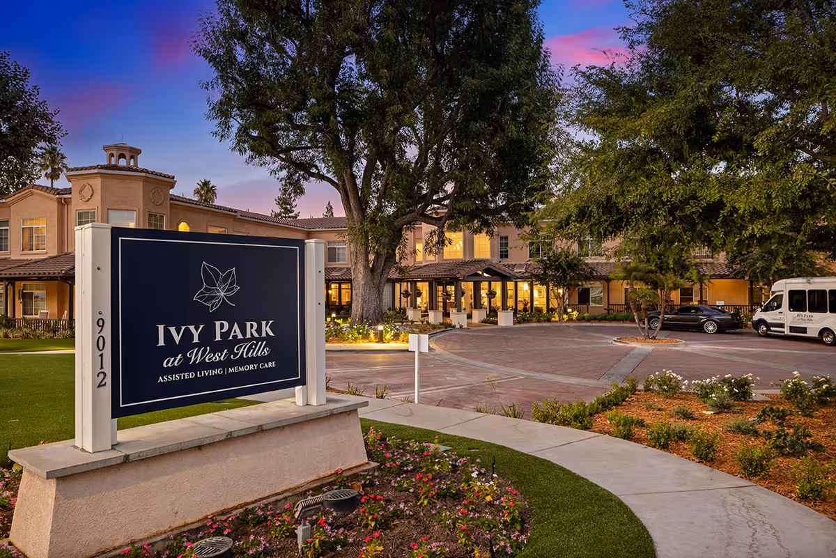 Exterior view of Ivy Park at West Hills assisted living and memory care facility during dusk, showing the main building with warm lights, a large tree, a circular driveway with parked vehicles, and a prominent sign with the facility's name and address surrounded by landscaped flowers and greenery.