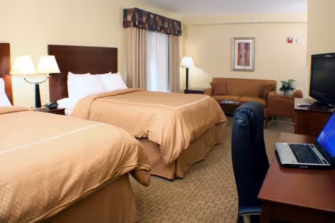 A hotel-style room with two double beds covered in beige bedspreads, each with white pillows. There are two bedside lamps on a nightstand between the beds. In the background, there is a window with curtains and a valance, a brown sofa, an armchair, a floor lamp, and a framed picture on the wall. In the foreground, there is a desk with a laptop and a black office chair.