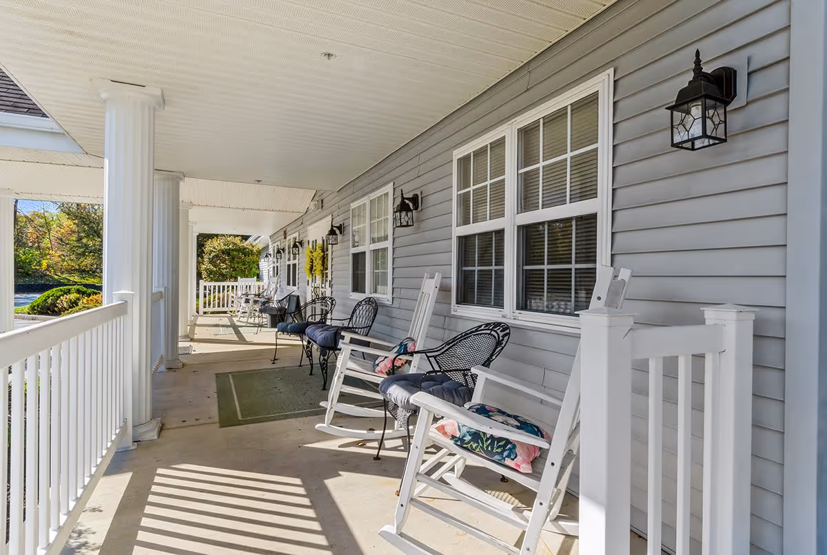 A covered porch area with white rocking chairs and black metal chairs with cushions lined up along the side of a gray building with white trim and windows. There are white columns supporting the roof and outdoor wall lanterns mounted on the building. The porch overlooks a landscaped area with greenery.