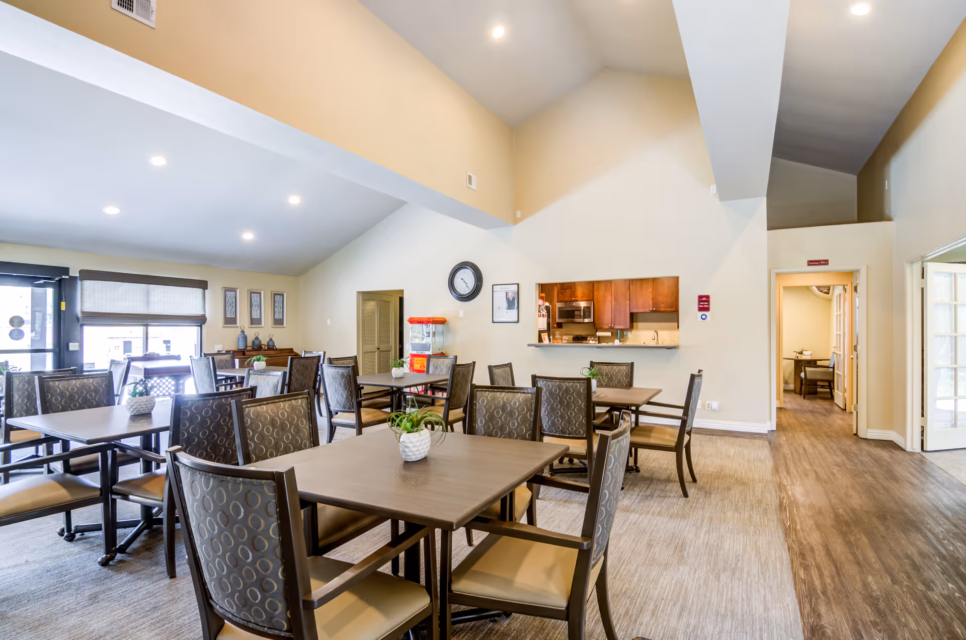 Interior view of a senior living facility dining area with multiple tables and chairs arranged neatly. The room has high ceilings and light-colored walls. There is a small kitchen area with wooden cabinets and a microwave visible through a serving window. A clock and framed pictures hang on the wall, and a popcorn machine is placed near the kitchen. The floor is covered with wood and carpet, and natural light comes through a window near the entrance.