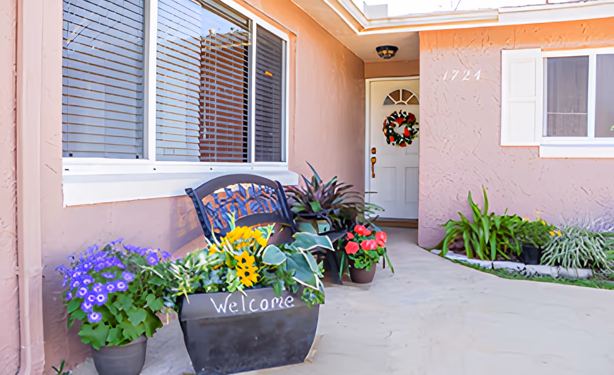 Front entrance of a residential building with a white door decorated with a holiday wreath. There are several potted plants and flowers, including purple, yellow, and red blooms, arranged along the walkway. A black planter with the word 'Welcome' written on it is placed near a decorative metal bench. The building exterior is painted light brown with white window frames and shutters.