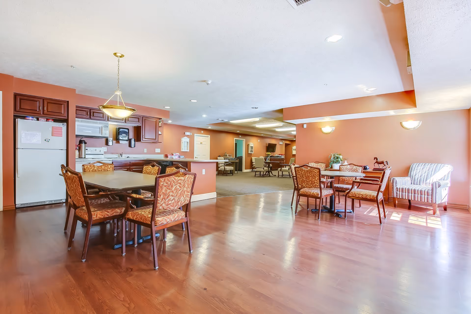 A spacious assisted living common area with a kitchen and dining tables. The kitchen has dark wood cabinets, a white refrigerator, microwave, and stove. There are two dining tables with patterned chairs on a wooden floor. The room is painted in warm tones with wall sconces and a hanging light fixture. In the background, there is a carpeted lounge area with additional seating and a television.