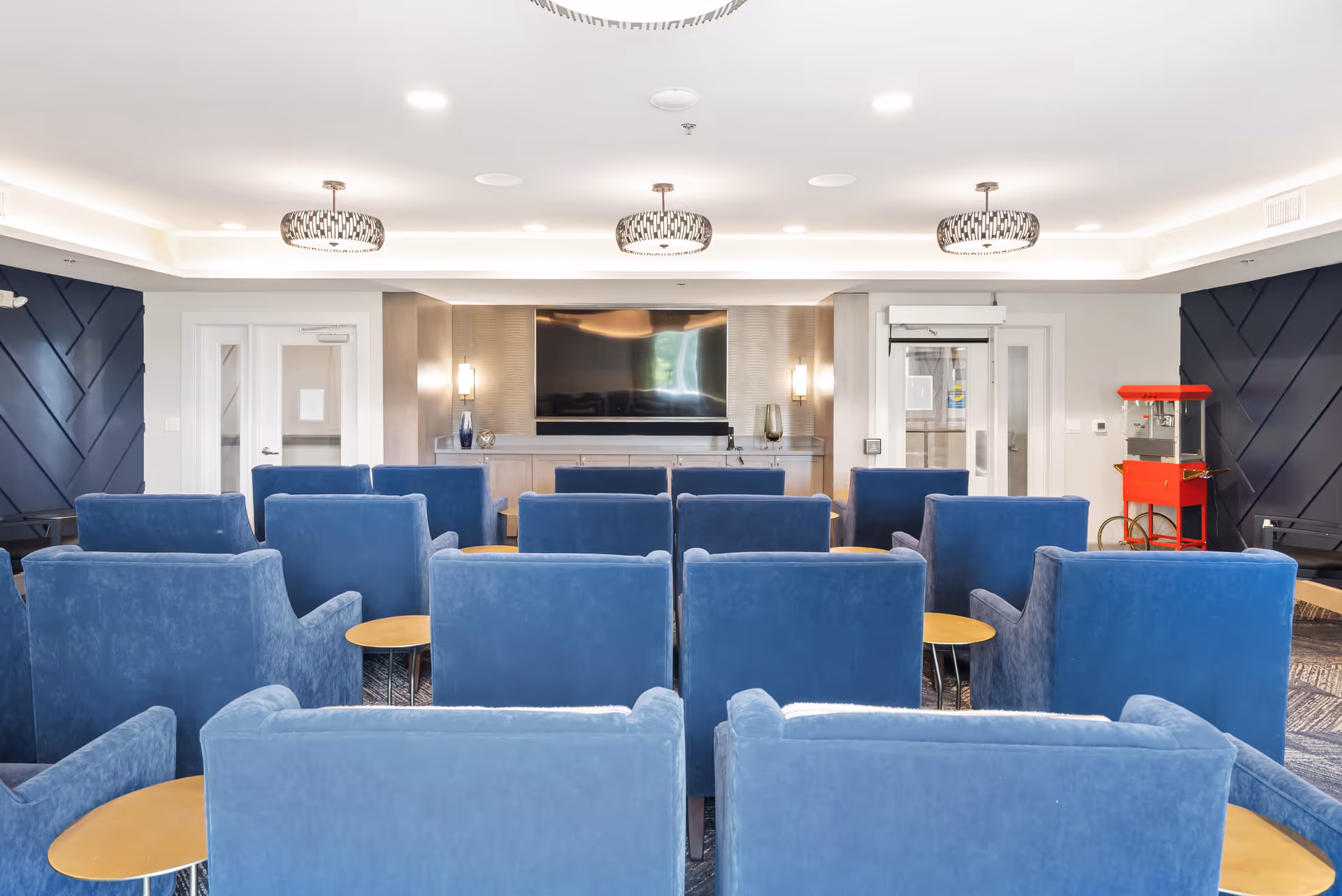 A modern common area with multiple rows of blue upholstered armchairs facing a large flat-screen TV mounted on the wall. The room features stylish ceiling lights, small round wooden side tables between chairs, dark blue accent walls with diagonal paneling, and a red popcorn machine on the right side near the wall.
