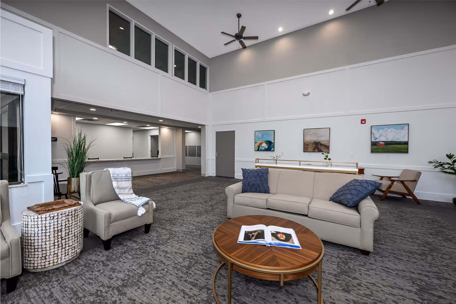 A spacious and modern senior living common area with high ceilings, neutral-colored walls, and carpeted floors. The room features a beige sofa with two blue pillows, two beige armchairs, a round wooden coffee table with an open book on it, and a side table with a decorative plant. Three framed paintings hang on the far wall, and a ceiling fan is mounted above. The area appears well-lit with recessed lighting and natural light from windows.