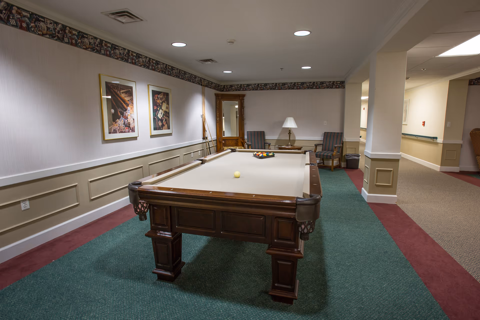 Interior room with a billiards table in the center, two framed pictures on the wall, two striped armchairs with a small table and lamp between them, and a wooden cabinet with a mirror in the corner. The room has green and maroon carpet and recessed ceiling lights.