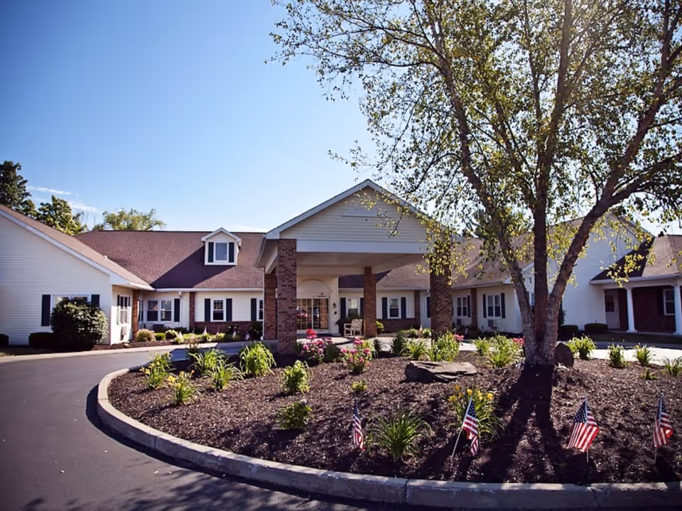 Exterior view of Keepsake Village at Greenpoint showing a single-story building with a covered entrance, landscaped garden with flowers and small American flags, and a large tree in the foreground under a clear blue sky.