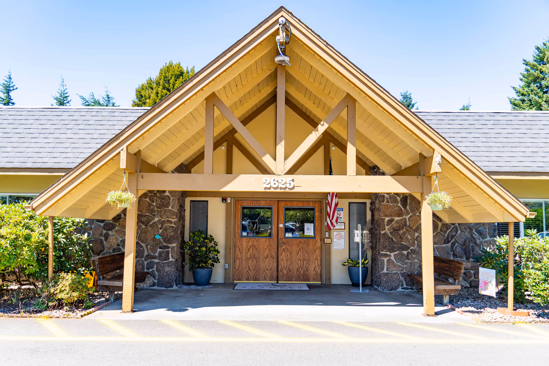 Front entrance of Avamere Rehabilitation of Coos Bay featuring a peaked wooden canopy supported by beams, stone walls on either side, double wooden doors with glass panels, potted plants, an American flag, and benches under the canopy.