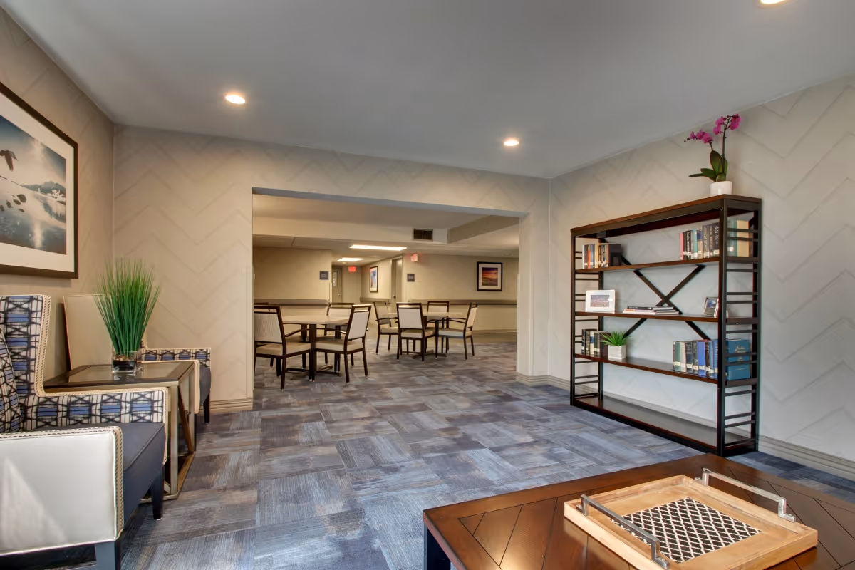 A spacious and well-lit common area in a senior living facility featuring a seating area with patterned armchairs and a small table with a plant, a wooden bookshelf with books and decorative items, and several tables with chairs in the background. The walls have a subtle chevron pattern, and framed artwork is displayed on the walls.