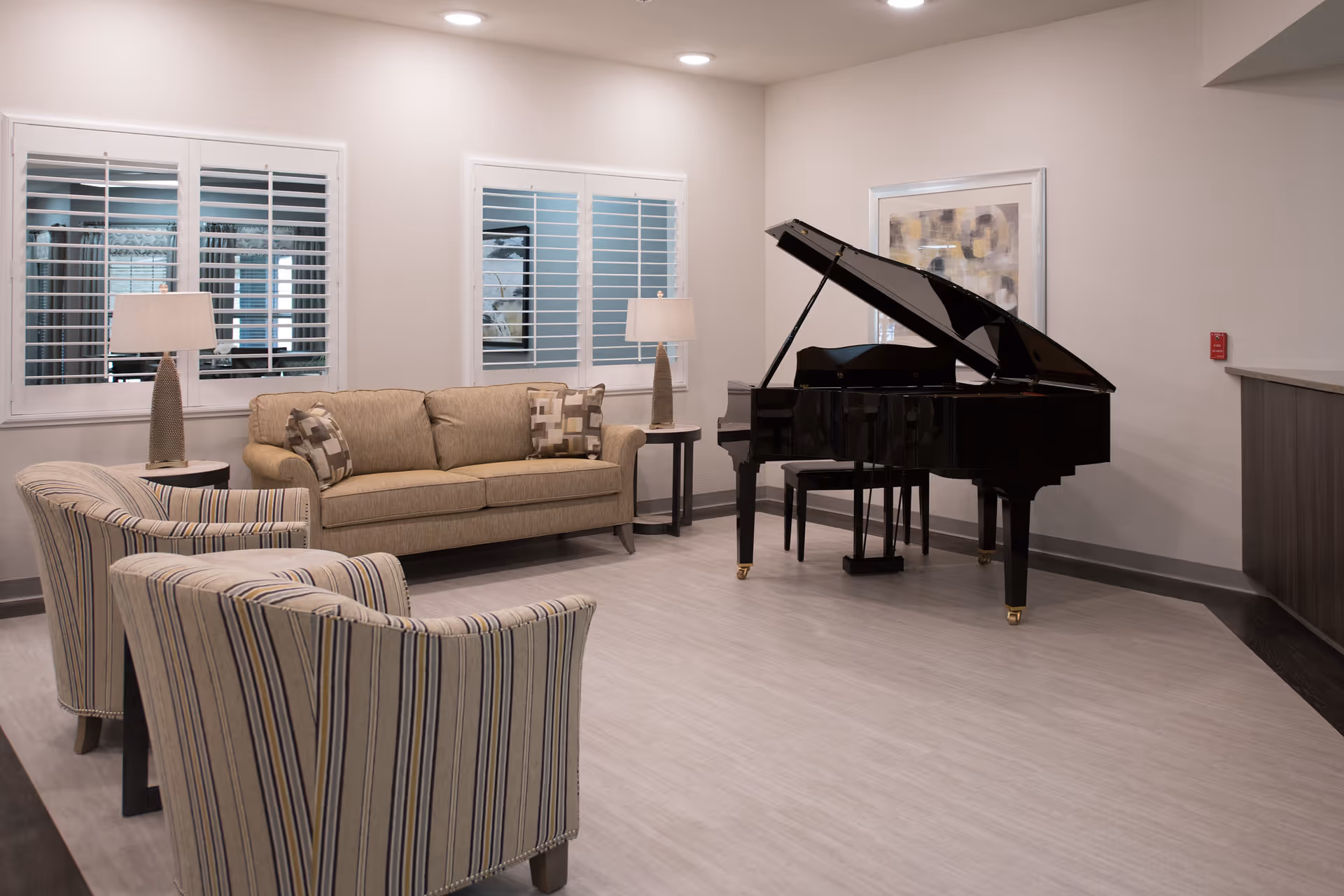A well-lit living room area with a beige sofa, two striped armchairs, two side tables with lamps, and a black grand piano. The walls are light-colored with two windows featuring white shutters and a framed abstract artwork above the piano.