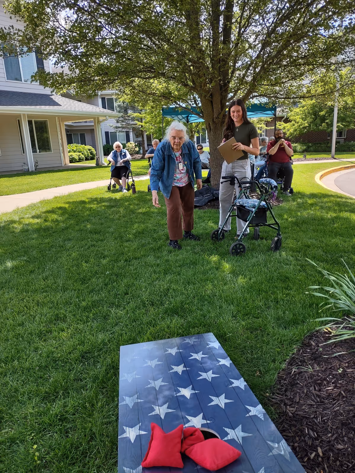 An elderly woman is playing a bean bag toss game on a grassy lawn under a large tree, with a younger woman holding a clipboard standing nearby. Several other elderly people are seated in the background near a building, enjoying the outdoor setting on a sunny day.