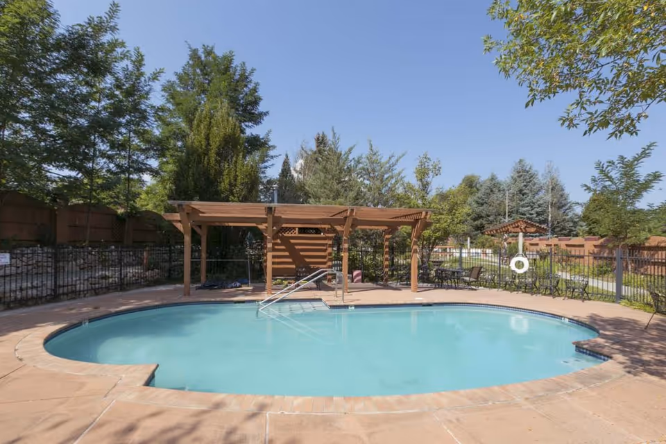 Outdoor kidney-shaped swimming pool with a wooden pergola, patio seating, and trees in the background.