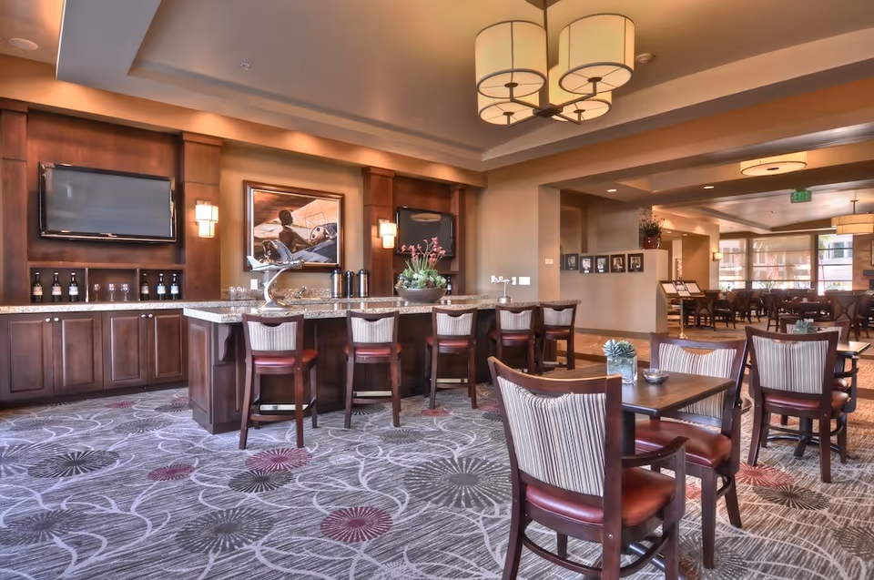 A spacious and well-lit dining area with a central island bar featuring six high chairs. The room has wooden cabinetry, two mounted flat-screen TVs, a decorative painting, and modern light fixtures. Several tables with chairs are arranged around the room, and the carpet has a floral pattern in shades of gray and purple.