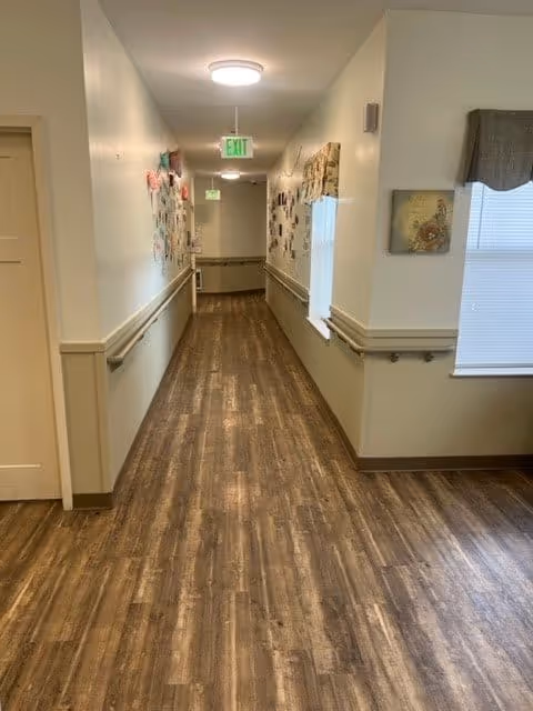 A long, narrow hallway with wood-patterned flooring and beige walls. Handrails run along both sides of the hallway. The left wall is decorated with various colorful papers and artwork. There is a window with a valance on the right side near the foreground, and an exit sign is visible at the far end of the hallway.