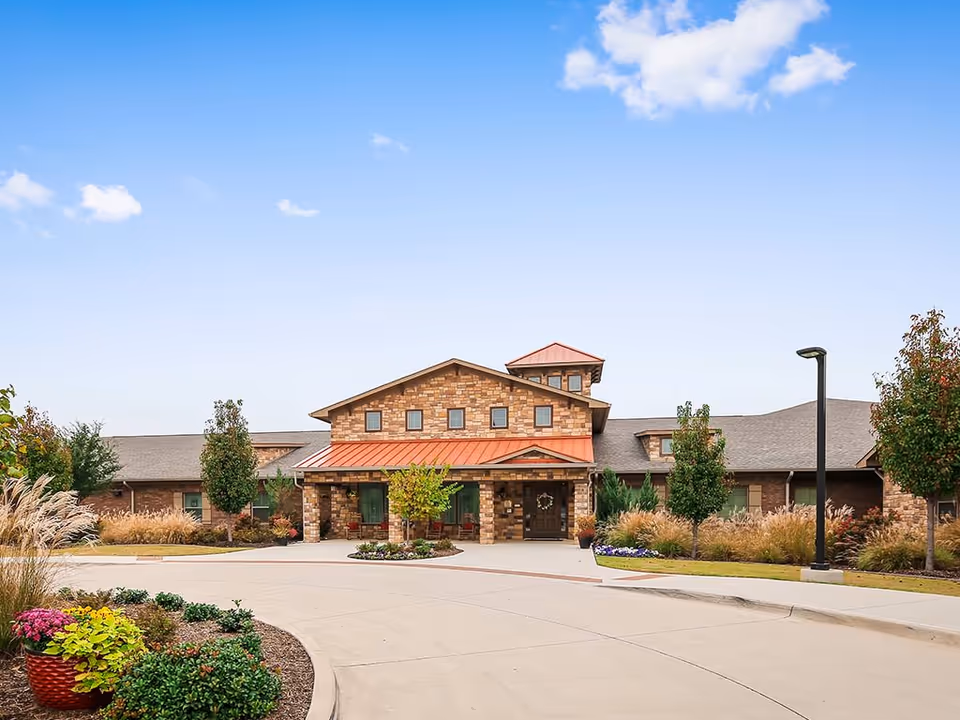 Stone-faced senior living facility entrance with a circular driveway, landscaped planters and a red metal awning under a blue sky.