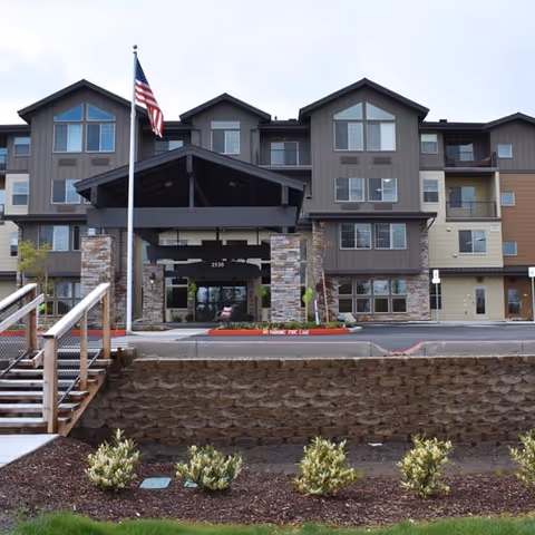 Front exterior view of a multi-story senior living facility building with a covered entrance supported by stone pillars, an American flag on a flagpole, and landscaped greenery in front.
