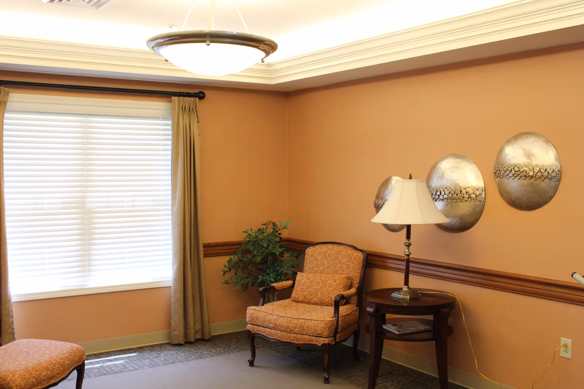 A cozy sitting area with an upholstered armchair, side table and lamp, window with blinds and curtains, and decorative wall plates.