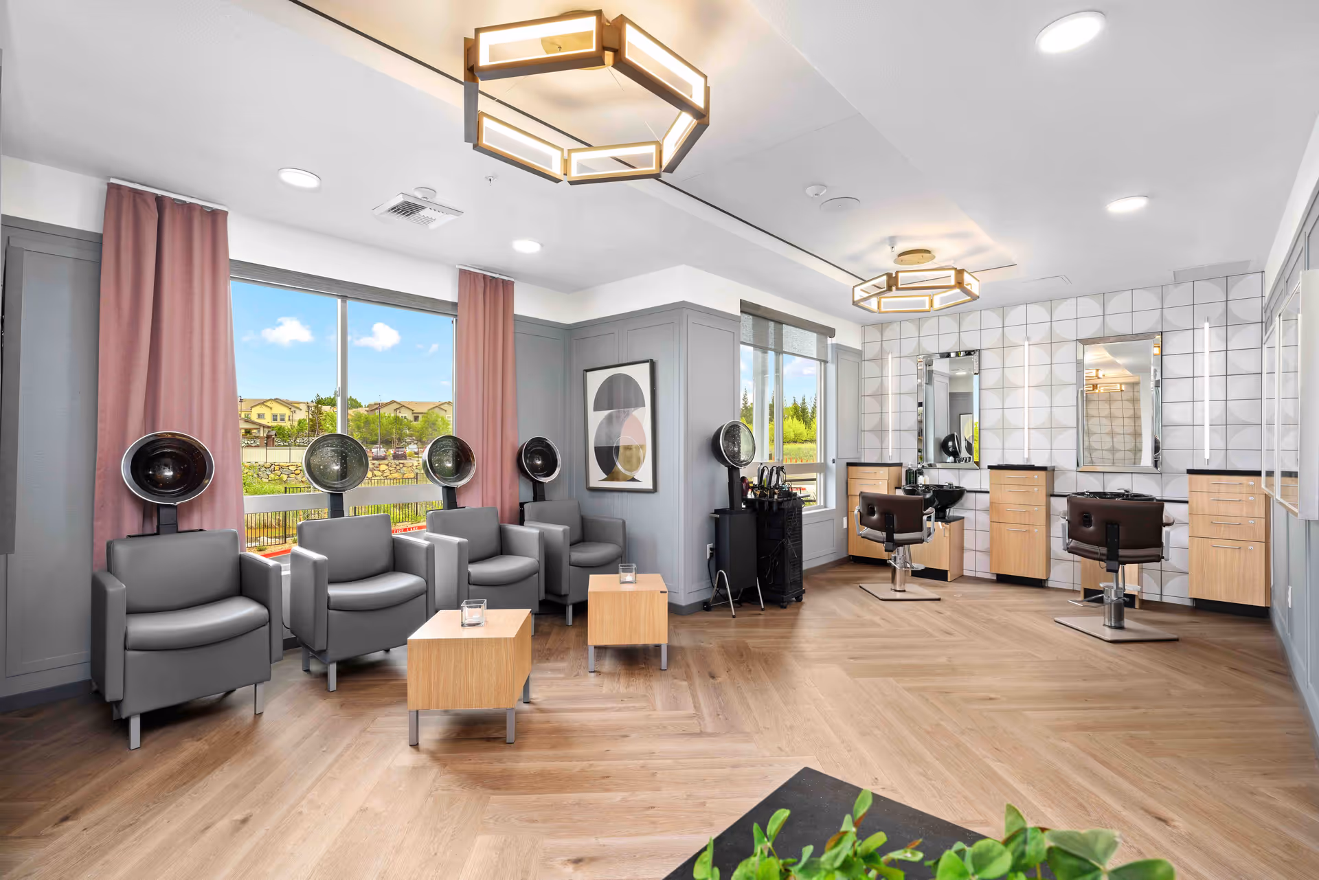 A bright and modern salon room with four gray salon chairs under hair dryers along the left wall, two wooden side tables in front of the chairs, and two styling stations with mirrors and brown salon chairs on the right side. Large windows with pink curtains let in natural light, and the room features wood flooring and contemporary ceiling lights.