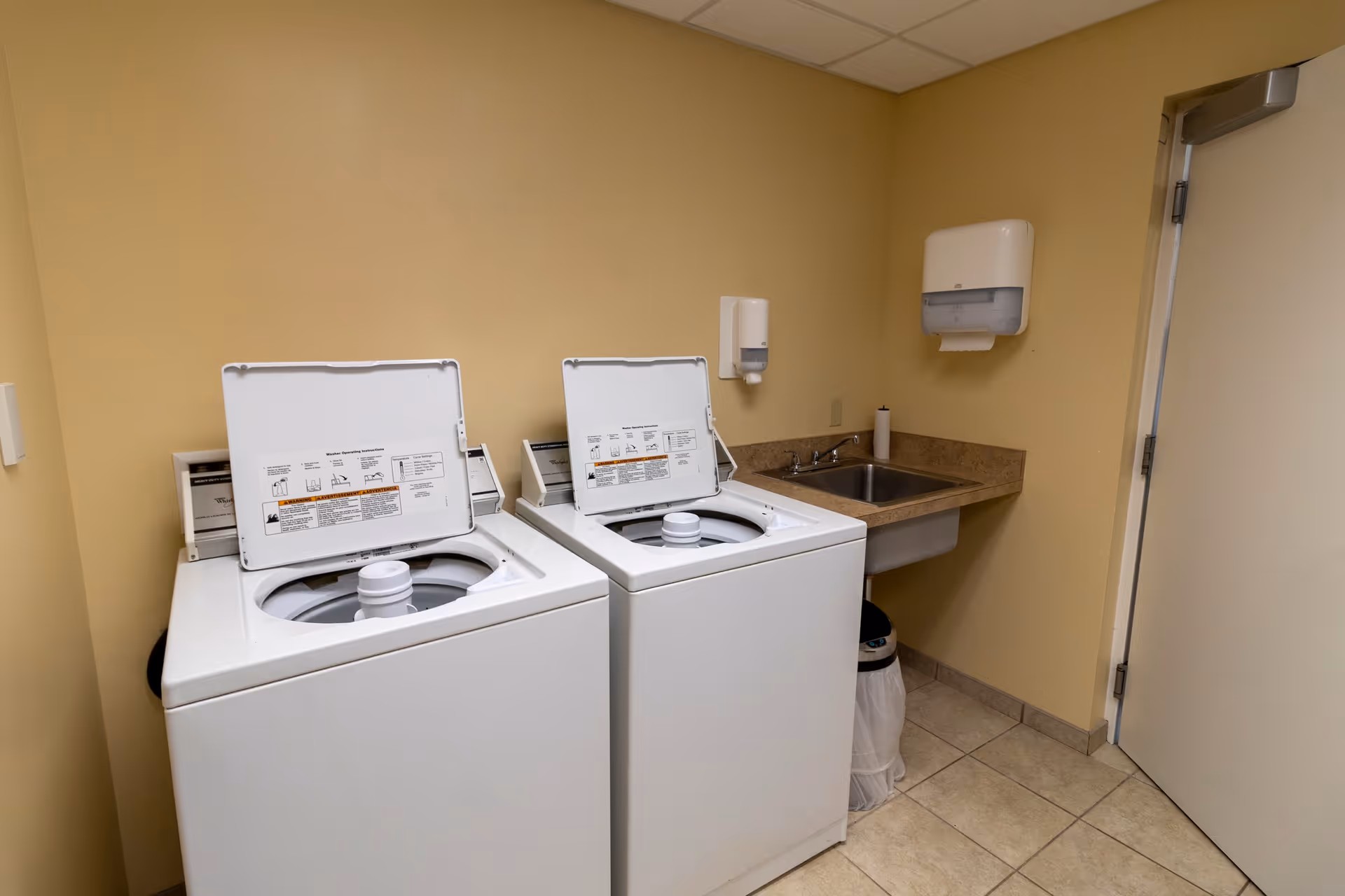 Laundry room with two top-loading washing machines, a sink with a faucet, a paper towel dispenser, a soap dispenser, and a trash can. The walls are painted beige and the floor is tiled.