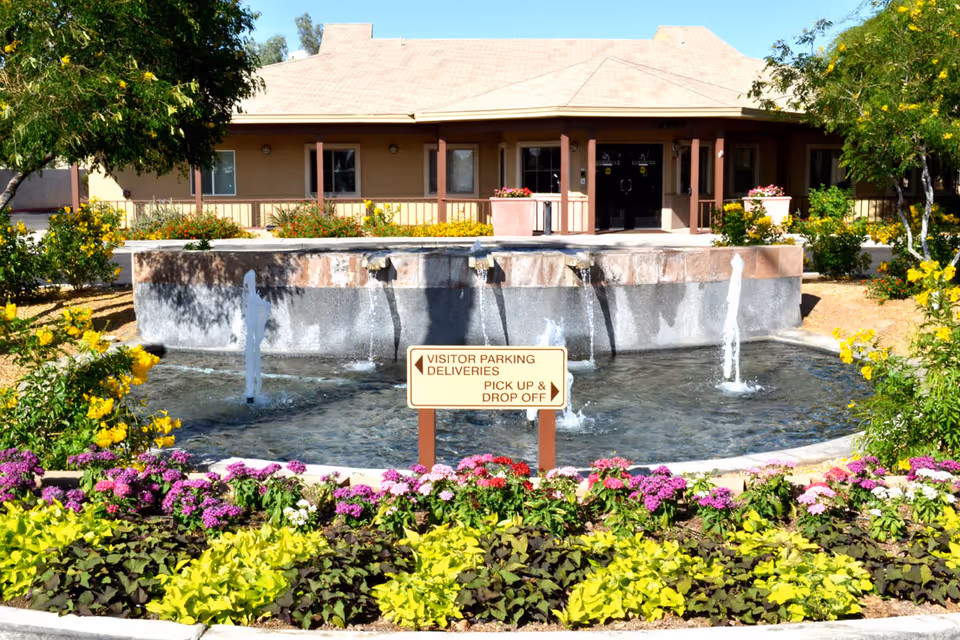 Outdoor view of a senior living facility named Sunshine Village featuring a water fountain surrounded by colorful flowers and greenery. The building entrance is visible in the background with a covered porch and glass doors. A sign in front of the fountain directs visitors to parking, deliveries, pick up, and drop off areas.
