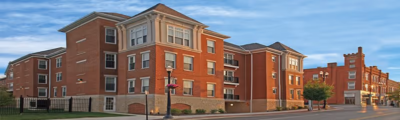 Exterior view of a multi-story brick building with multiple windows and a streetlamp with hanging flower baskets in front, under a partly cloudy sky.