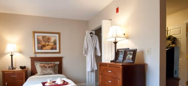 A cozy bedroom in a senior living facility featuring a single bed with a decorative pillow and a tray with a cup and napkin on it. There is a wooden nightstand with a lamp and a clock, a framed landscape painting on the wall, a wooden dresser with framed photos and a lamp, and a white robe hanging on the open closet door.
